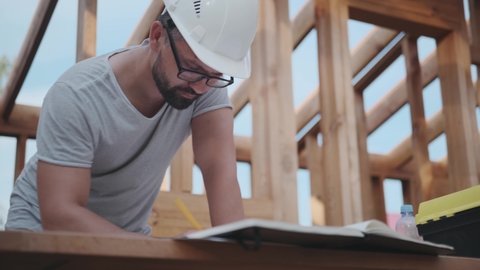 The architect inspects the structure and makes notes on the project. A man in a helmet is a designer working on a project. Home construction: designer engineer on building structure background
