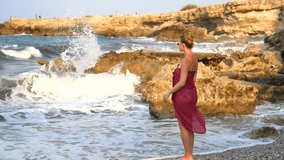Young slender beautiful girl in swimsuit and burgundy pareo stands on seashore and looks at sea and horizon, relaxes, meditates. Summer holidays, sense of joy. Slow motion video. Waves crash on rocks.