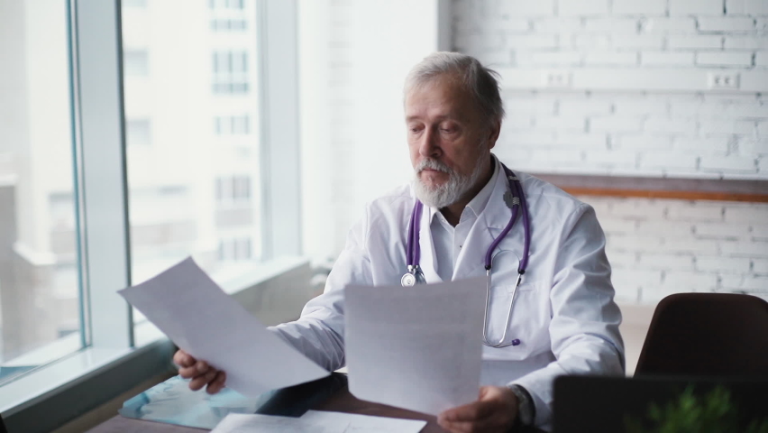 Experienced caucasian adult doctor with white beard and grey hair are siting at desk and studying patients reports, against large window in modern office room of the hospital. Shooting in slow motion. | Shutterstock HD Video #1058819602