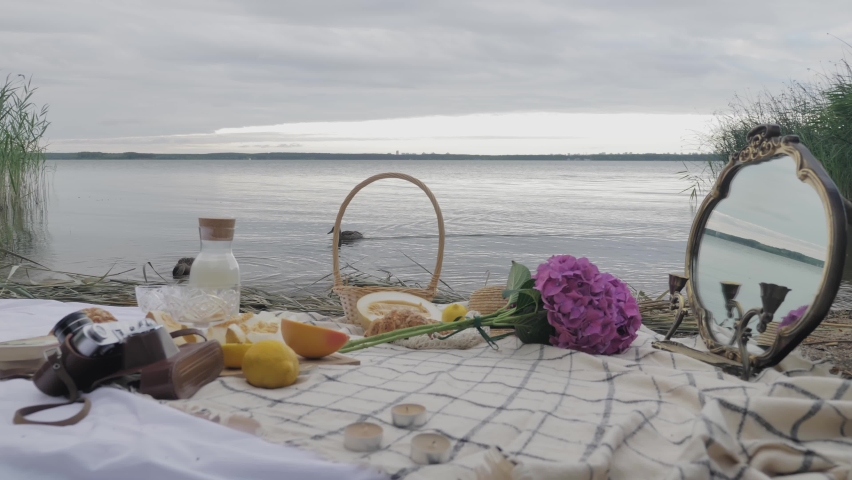 Picnic against the background of the water on which the ducks swim. On the veil there are flowers, a basket of fruit, a film camera, a carafe with milk, a melon, a vintage mirror.