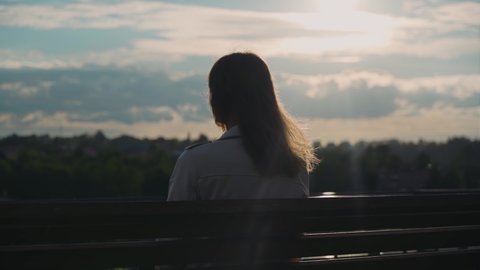 A long-haired girl sits on a bench against the backdrop of the evening sky