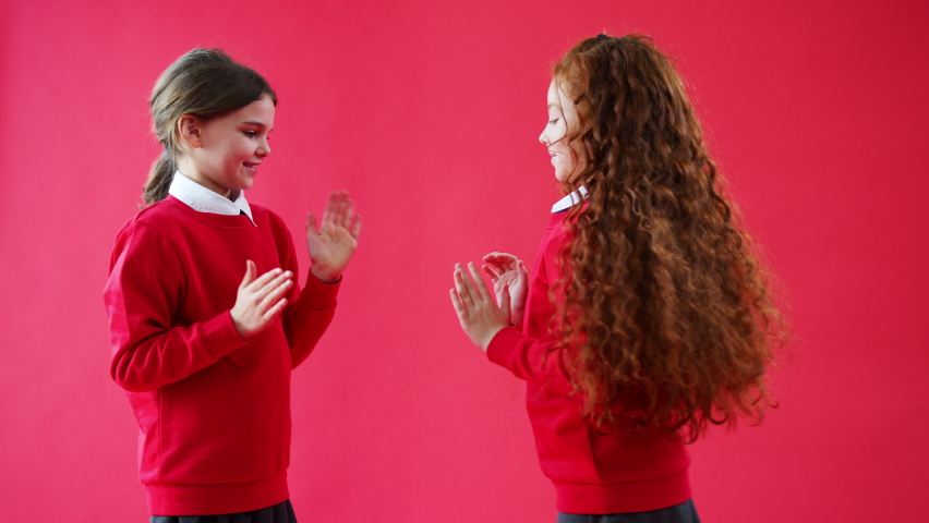 Two Female Elementary School Pupils In Uniform Playing Clapping Game Against Red Studio Background