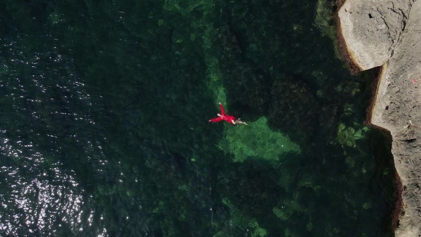 woman in a red long dress is lying on her back bathing in the green transparent water, of the sea next to a rock in summer