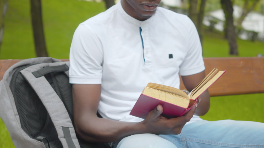 Unrecognizable African American college student reading out loud in sunny park. Concentrated young man sitting on bench with book and backpack. Lifestyle and hobby concept.