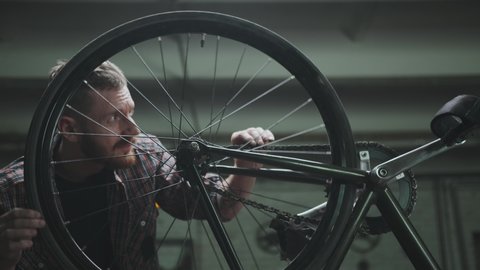 A male cyclist in a plaid shirt repairs a bicycle in a garage, turns a wheel and checks it. Vehicle repair: bike service center concept. Medium shot.