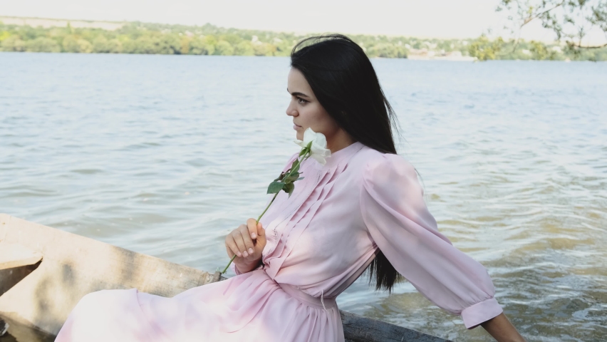 A girl dressed in a pink dress with a white flower in her hand sits in a boat and admires the view.