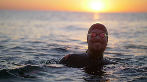 Smiling male swimmer athlete in swimming cap and goggles in the sea at sunrise, having fun, happy, satisfied sportsman. High quality 4k footage