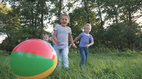 A happy family in the open air in the park spends the weekend. Children run after a colored inflatable ball in a crowd overtaking each other. Little kids play on the grass with the ball. Teamwork for