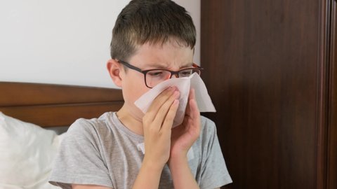 A boy with flu and fever lying in bed and blowing his nose with a paper tissue, seasonal viral diseases concept.