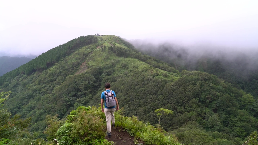 Male hiker standing on top of epic Mountain lookout, looking towards hills with forest and rolling clouds