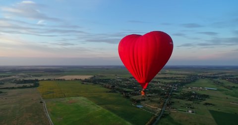 Flight of a red airship. Beautiful hot air balloon in heart shape travelling over the green fields in the morning. Aerostat with basket in the sky.