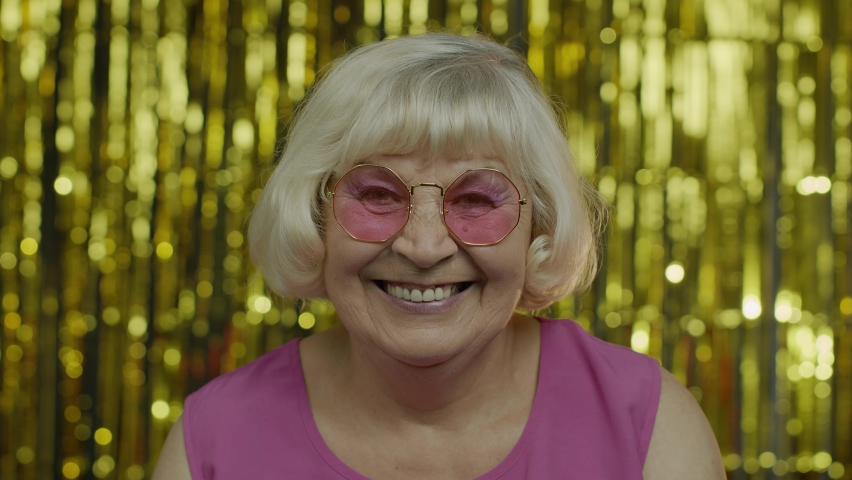 Portrait of positive senior old blond woman in sunglasses looking around, enjoying, smiling, satisfied with life, good mood, success. Elderly stylish lady grandma on golden background. 6k resolution | Shutterstock HD Video #1058847799