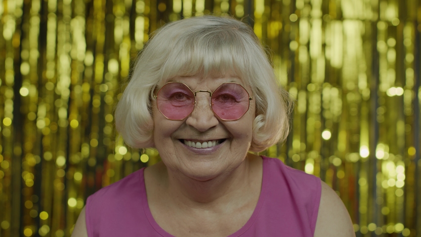 Portrait of positive senior old blond woman in sunglasses enjoying and smiling, satisfied with life, good mood, success. Elderly stylish lady grandma on golden background in studio. 6k footage | Shutterstock HD Video #1058847805