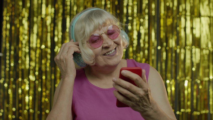 Stylish happy senior old woman in pink blouse holding cell phone, listening to energetic music in headphones, dancing enjoying favorite song. Elderly lady grandma on golden background. 6k resolution | Shutterstock HD Video #1058847847