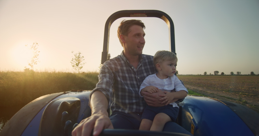 Authentic shot of happy farmer father is driving a tractor with his little son and showing a family agricultural possession in a sunny day. Concept: agriculture, farming, family, parenting, childhood Royalty-Free Stock Footage #1058850883
