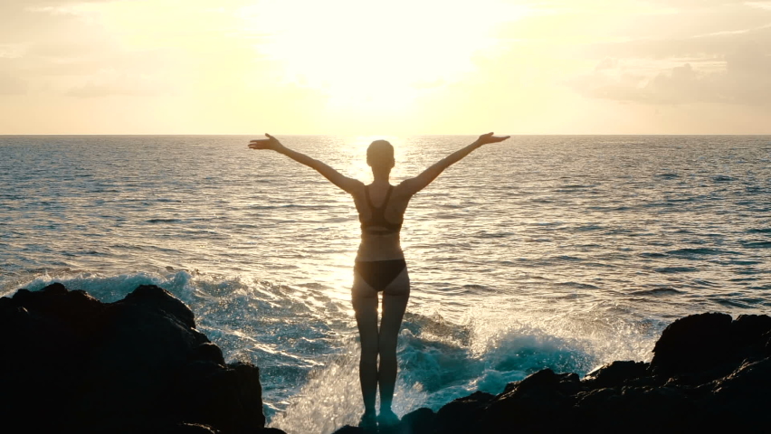 Silhouette woman at sunset raising arms in air and observing ocean waves crashing on rocks and spraying. Concept of explosion of emotions, joy, happiness in life. Traveling around world, slow motion.  Royalty-Free Stock Footage #1058851294
