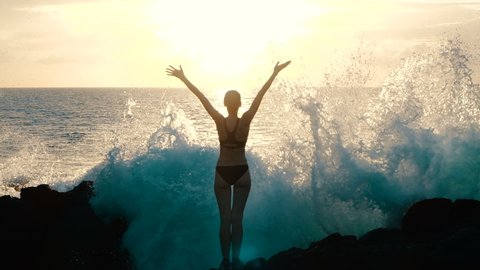 Silhouette woman at sunset raising arms in air and observing ocean waves crashing on rocks and spraying. Concept of explosion of emotions, joy, happiness in life. Traveling around world, slow motion. 