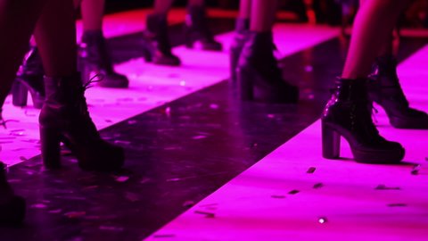 Men and women in stage shoes perform a dance on stage, bouncing and moving in sync. A close up of legs and shoes from the bottom. 