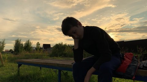 A lonely sad boy sitting outdoors on a bench at sunset, unhappy depressed child with discrimination and bullying problems.