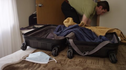 Man throws clothes from a closet into a suitcase laying on a bed with a face mask