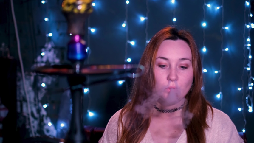 Beautiful woman blows smoke in night club or hookah or shisha bar. Media. Red haired young woman exhaling white smoke through her mouth and nose.