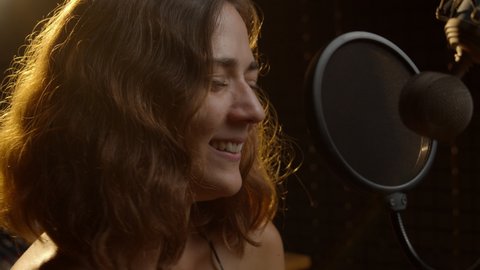 Song recording in the music studio. Hispanic woman singing close to the microphone. Curly brunette female person performs songs. 