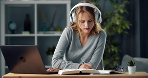 Pretty young woman with blond hair using wireless headphones and laptop for studying at home. Female student preparing for exams with modern gadgets.