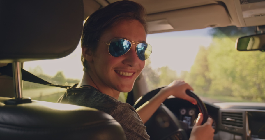 Young happy guy in the car looking to the camera. Handsome young smiling man in sunglasses is sitting in a car at sunset. Sun