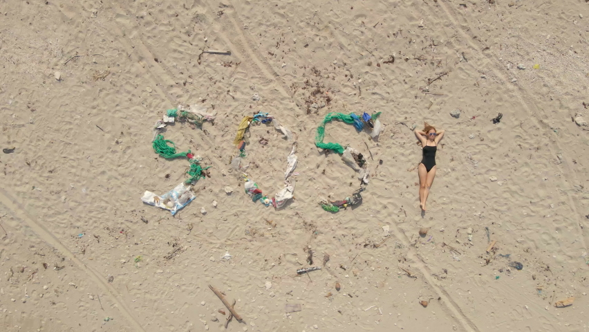 Aerial shot of the sign SOS made of trash on a beach. Woman lays by the sign simbolizing an exclamation mark. Ecologic disaster concept. Plastic pollution concept