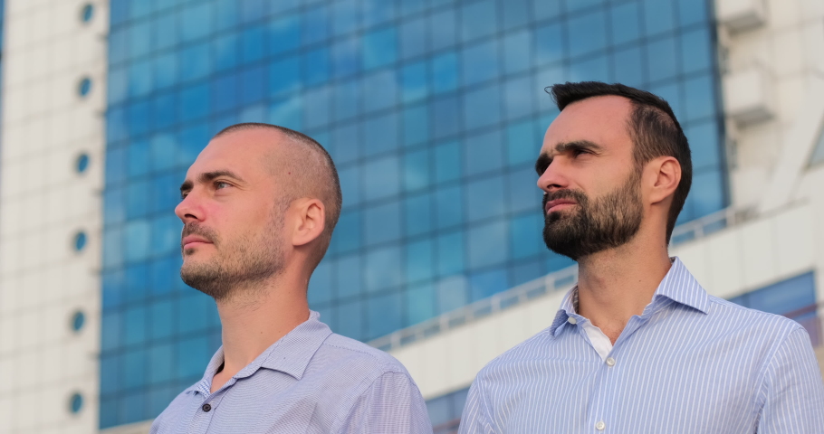 Two men put on white safety helmets in front of a building with blue windows, engineers look in one direction.