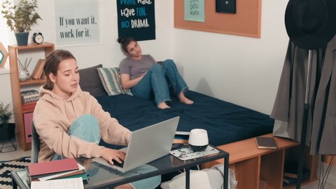 Tracking shot of young female college student sitting at desk in her dorm room and doing assignment on laptop. She is asking question from her cheerful roommate relaxing on bed