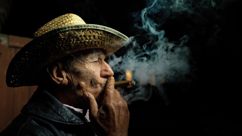The farmer is resting in the evening over a good cigar. An elderly farmer in a straw hat smokes a cigar on a farm near a barn. Confident Senior reflects in cigarette smoke. An old cowboy at the ranch. | Shutterstock HD Video #1058877004