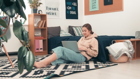 Handheld dolly-in shot of cheerful girl sitting on floor in cozy college dormitory and typing on laptop while doing her assignment
