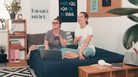 Handheld shot of happy young women sitting on bed in cozy bedroom or college dormitory and dancing and singing to music while looking at camera