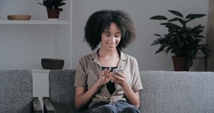 Smiling resting Afro American millennial woman hold smart phone in hand watching social media stories video sit on sofa in living room. Happy mixed race lady using mobile apps, laughing look at camera