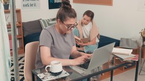 Tracking shot of young female student in glasses sitting at desk in dormitory and typing on laptop. Her female roommate sitting on bed and reading textbook - Powered by Shutterstock - Get 15% off with code: PIKWIZARD15