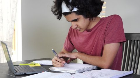 Medium shot of teen boy of Persian ethnicity doing remote school in times of Covid-19, taking math notes from a virtual lesson on his laptop. Matching close up available.