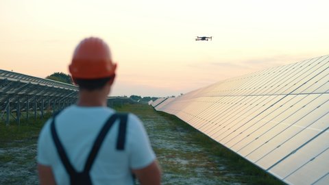 Engineer in hardhat holding tablet computer operating flying drone in solar plant. Photovoltaic solar panel installation. Solar array. New technologies.