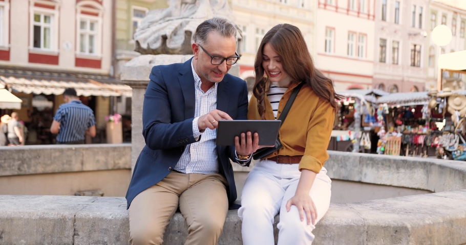 Handsome middle-aged man in glasses sitting on fountain with pretty young female and showing something on tablet device. Outside. Caucasian father and his beautiful adult daughter using gadget computer.