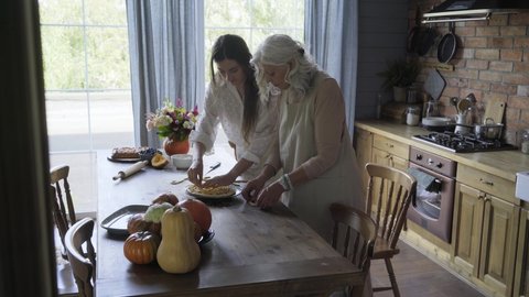 aged lady with loose grey hair prepares pumpkin pie with daughter help on wooden table near fresh fruit against large plastic kitchen windows 스톡 비디오