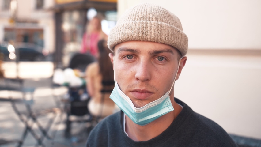 A portrait of a young caucasian man takes off a medical mask. A man sits in a cafe outside