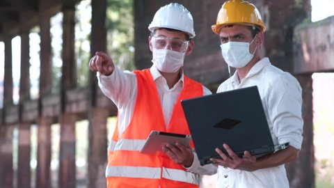 Workers in helmets and medical masks with a tablet and laptop discuss reconstruction plans farms. Overhaul and maintenance of buildings. Making a decision on implementation of repair work.