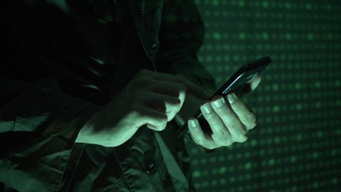 Hacker using a mobile phone to hack the system. He is standing in the dark with a matrix-like background.