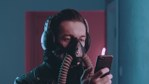 Cyberpunk portrait of brutal man in respirator enjoys listening to music with headphones on his smartphone in neon light. Male in gas mask listening to music on blue and pink neon light background.