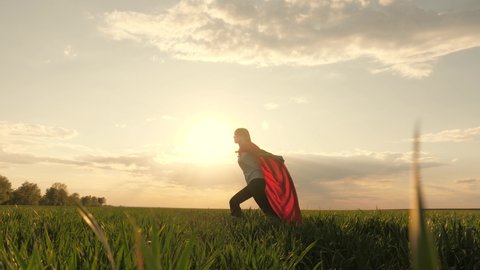 happy superhero girl, runs on green field in red cloak, cloak flutters in wind. child plays and dreams. Slow motion. teenager dreams of becoming superhero. young girl in red cloak, dream expression.