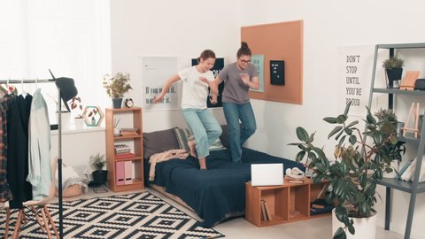 Tracking shot of young female friends laughing and having fun while dancing on bed in cozy bedroom or college dorm room