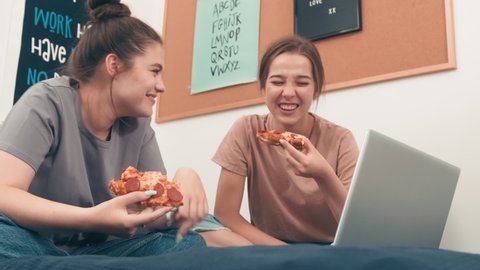 Low angle handheld shot of happy young women sitting cross-legged on bed and laughing while eating pizza and watching funny movie on laptop