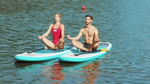Close up at sunlight man and woman is sitting in a lotus pose on a board during practicing yoga or meditation surfboard soak water swimsuit freedom active close up slow motion
