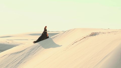 beautiful young woman in luxurious black silk dress walks in the desert. fabric of long train flutters in wind in slow motion 4k video footage. Background white sand dunes sunset. Happy girl tourist 