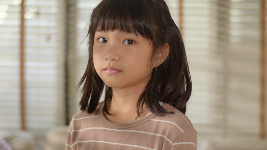Portrait of Funny Cute Asian Little Girl looking at the camera posing and smiling with funny, positive, and happy emotion while standing in the living room at home. Slow-motion and Sensitive Focus. Royalty-Free Stock Footage #1058928836